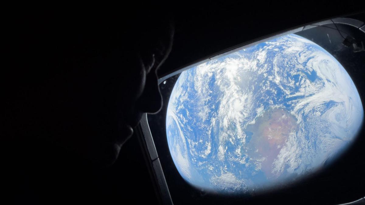 A person’s silhouette looks out a spacecraft window at Earth—a blue and white sphere against the darkness of space—reflecting the hope inspired by Artemis II’s journey to the moon.