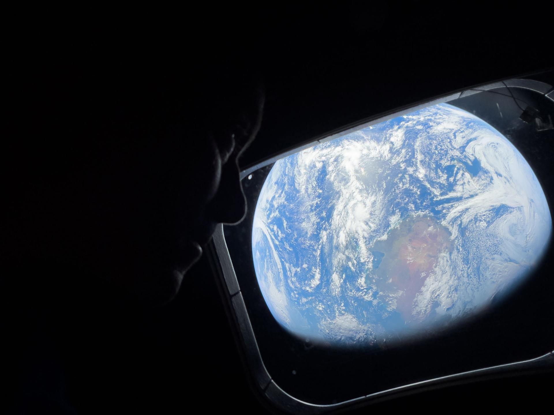 A person’s silhouette looks out a spacecraft window at Earth—a blue and white sphere against the darkness of space—reflecting the hope inspired by Artemis II’s journey to the moon.