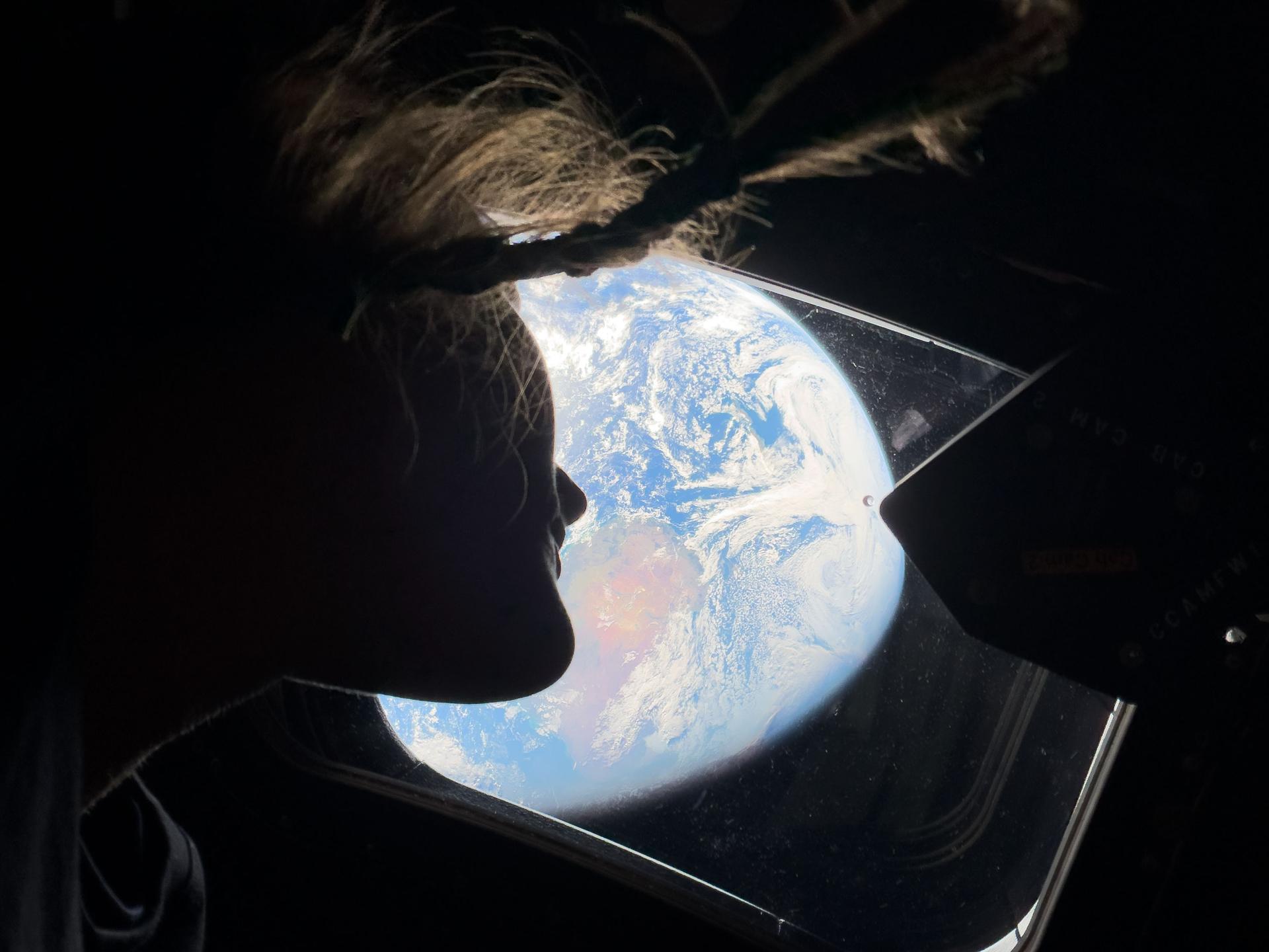 A person with hair floating upwards looks out of a spacecraft window at Earth, filled with hope as clouds and oceans shimmer below&mdash;a view reminiscent of the Artemis II mission&rsquo;s journey.