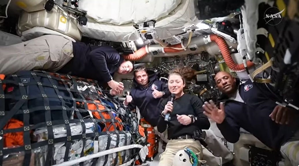 Four Artemis II astronauts in a spacecraft cabin, surrounded by equipment, smile and give thumbs up, with one holding a microphone&mdash;radiating hope as they prepare for their historic journey to the moon and look forward to seeing Earth from afar.
