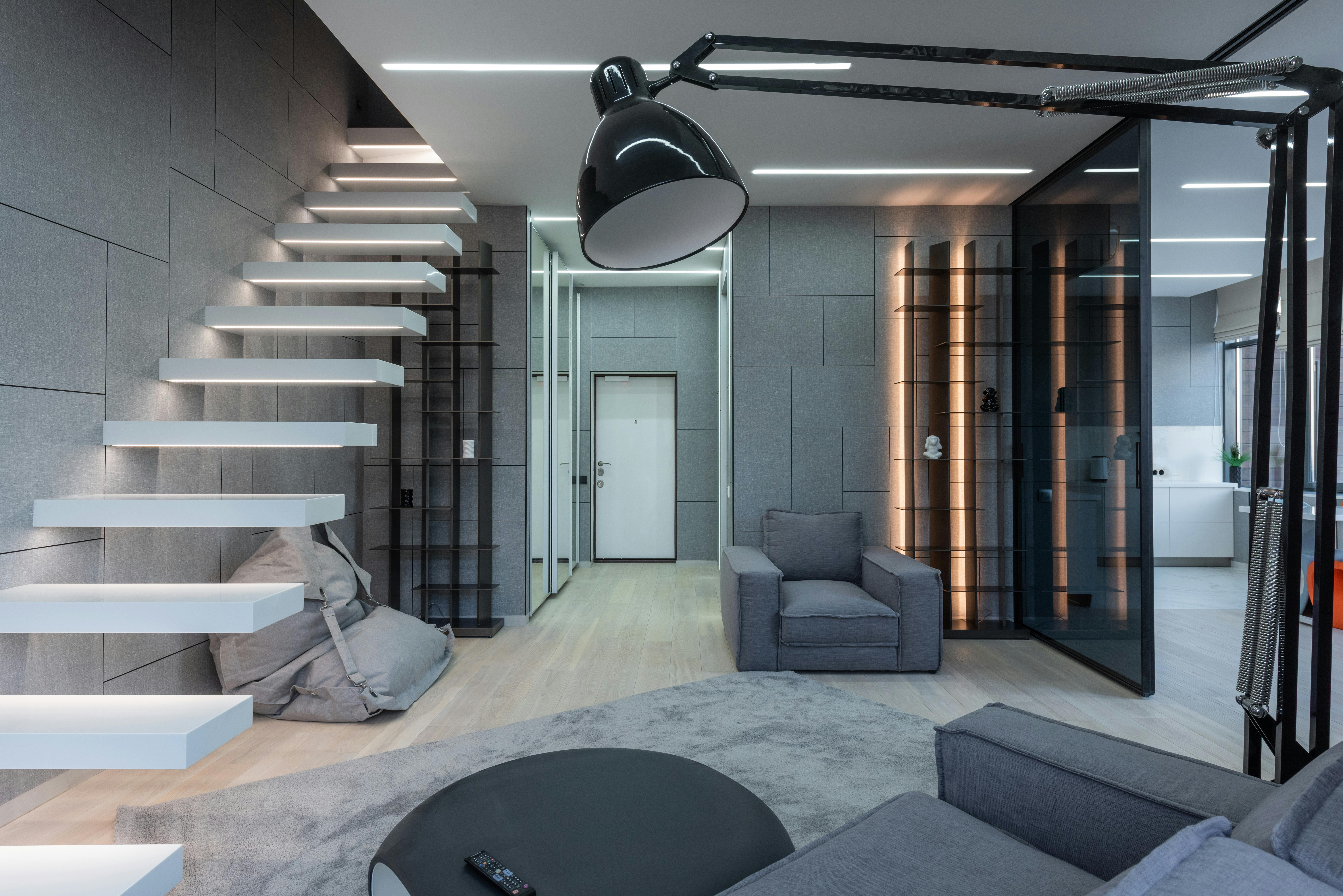Modern living room with gray furniture, floating staircase, wall-mounted shelves, large floor lamp, glass doors, and neutral decor.