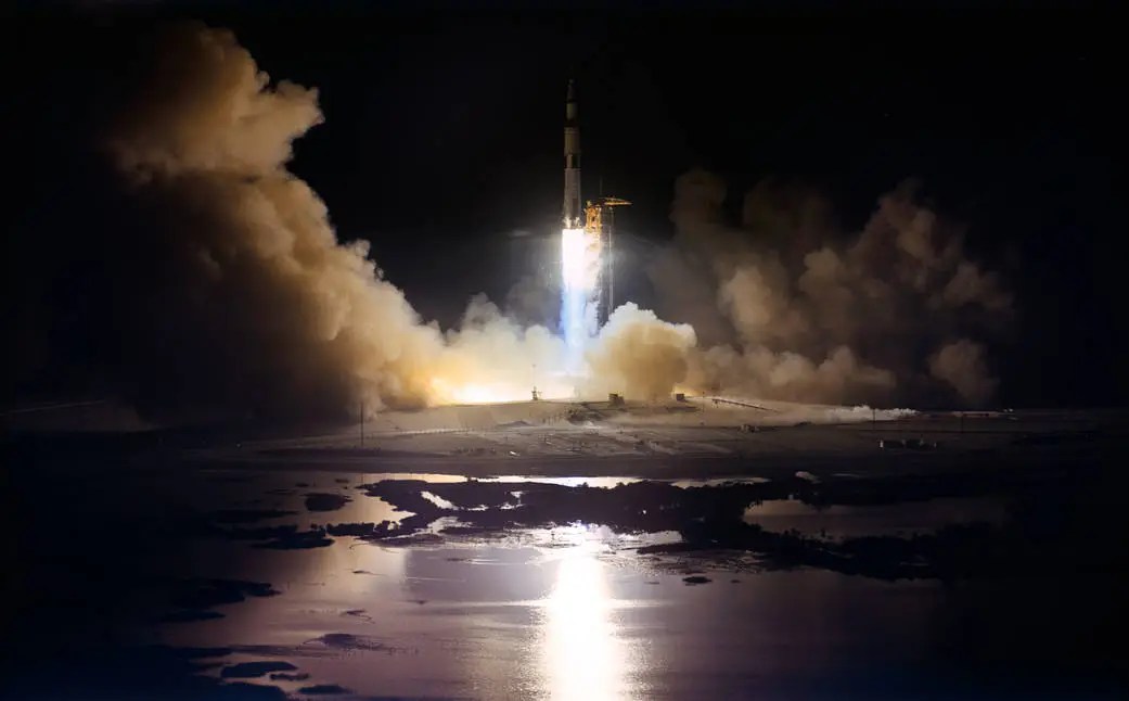 A rocket launches at night, emitting bright flames and smoke, its reflection visible in the water below&mdash;a powerful symbol of hope as Artemis II sets out on its journey from Earth to the moon.