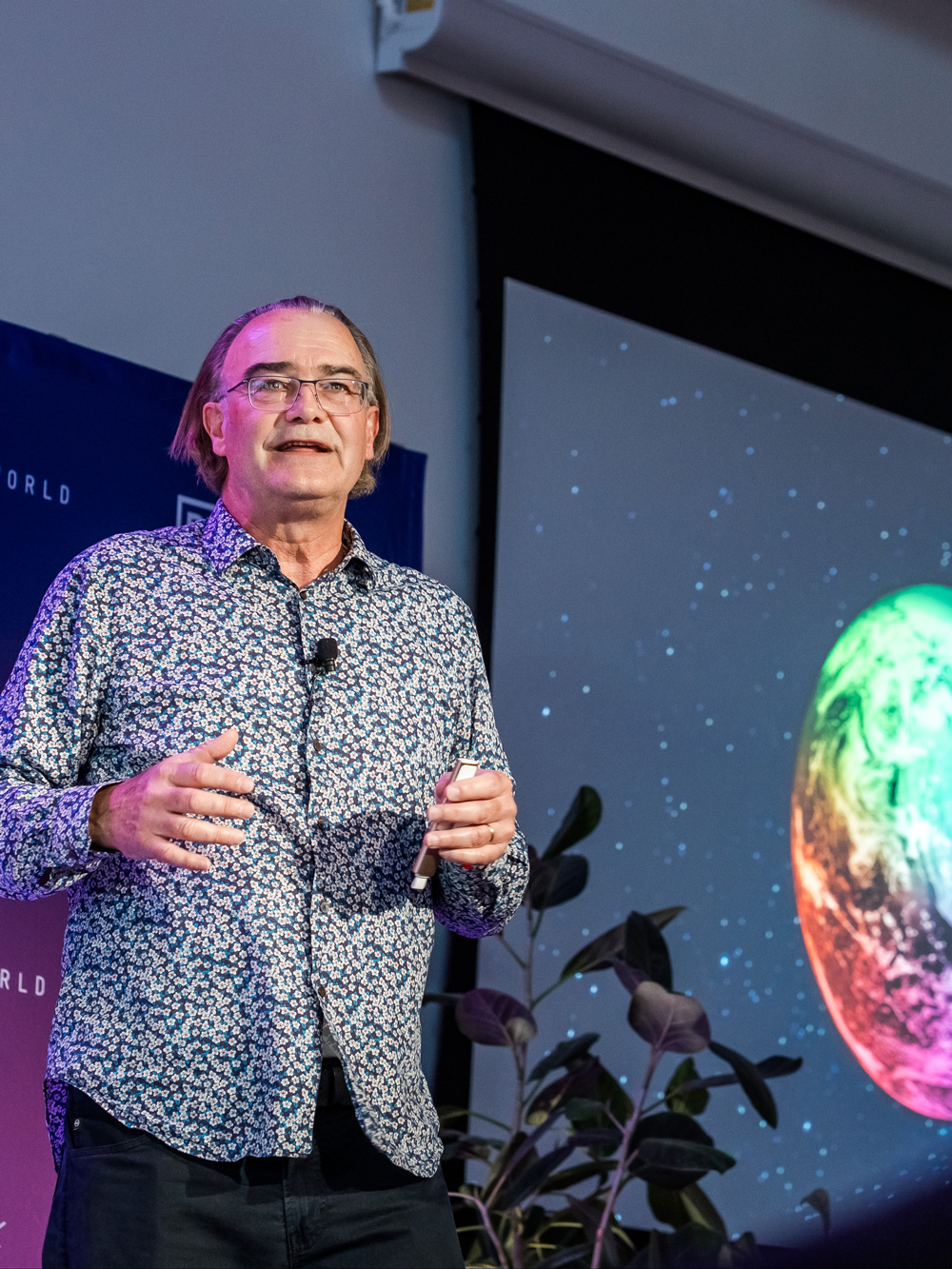 A man in a patterned shirt stands and speaks on stage next to a screen displaying a colorful image of a planet or Earth against a starry background.
