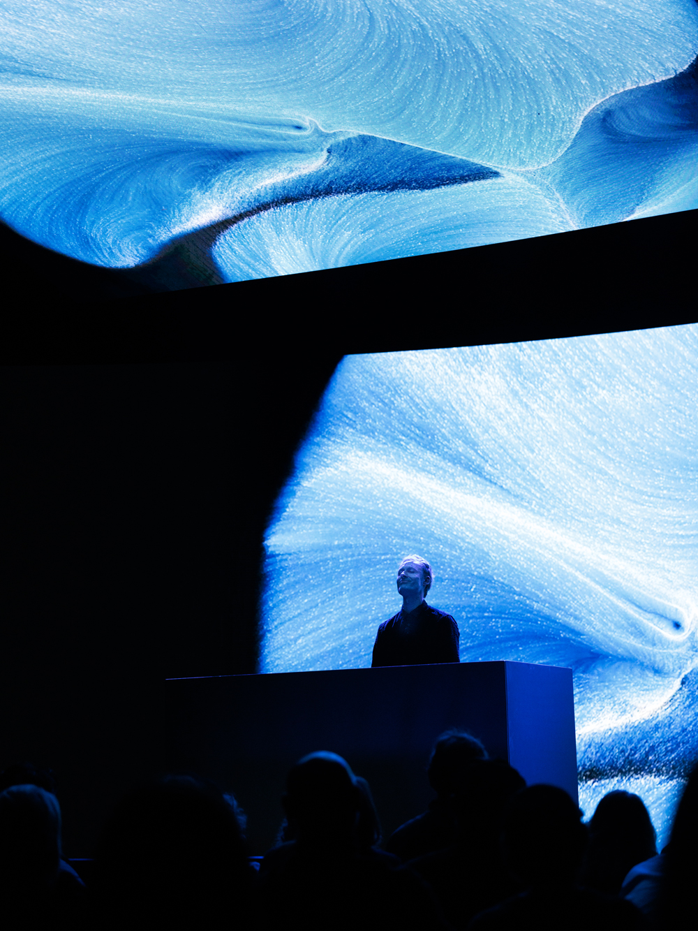 A person stands at a podium on stage with abstract blue and white digital art projected on large screens behind and above them.