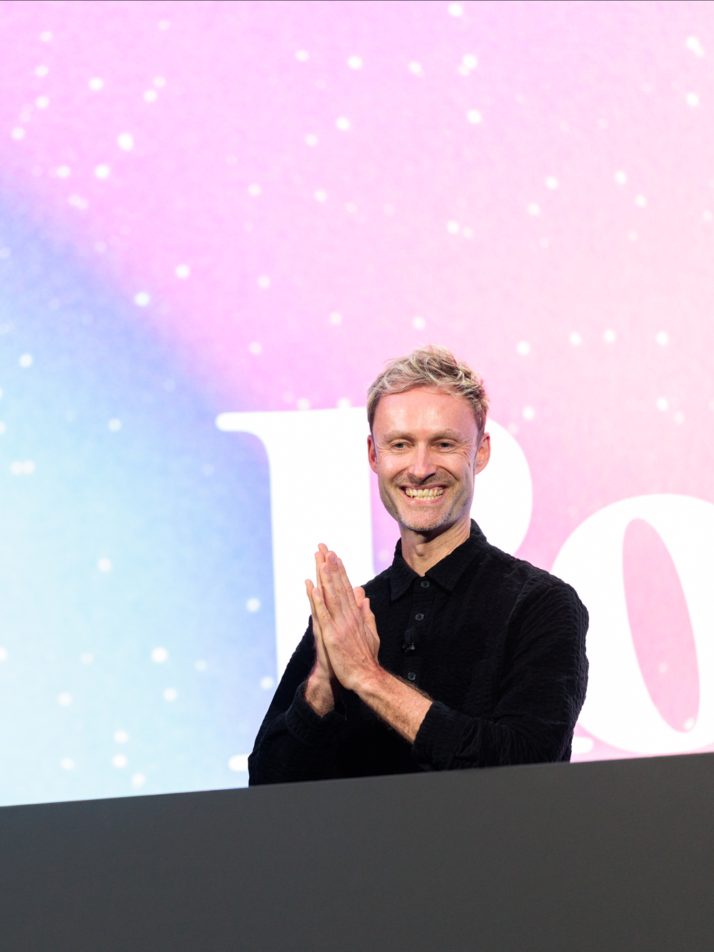 A man with blond hair and a black shirt stands in front of a colorful background, smiling and clasping his hands together.