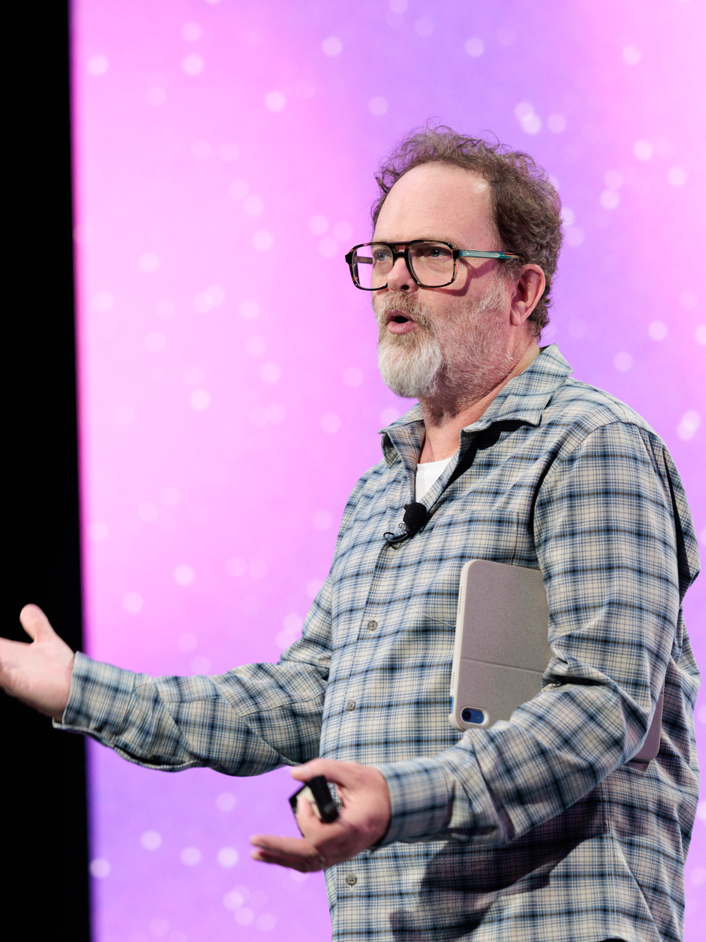 A man with glasses and a beard speaks on stage, holding a tablet, with a microphone clipped to his shirt and a purple background behind him.