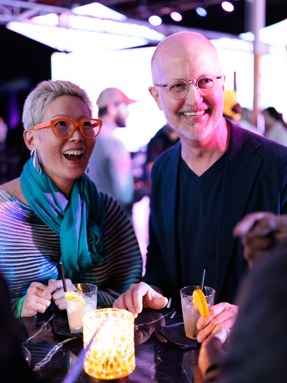 Two people with drinks in hand stand at a table, smiling and talking, under colorful lights at a lively indoor event.