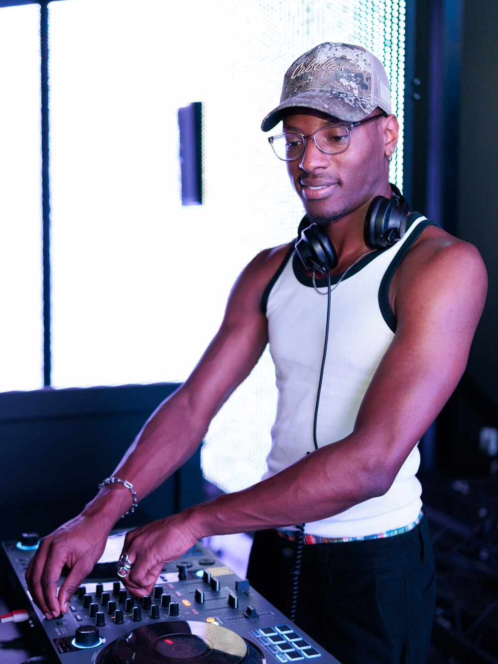 A person wearing a tank top, glasses, and a cap is standing at a DJ mixer with headphones around their neck, adjusting the controls.