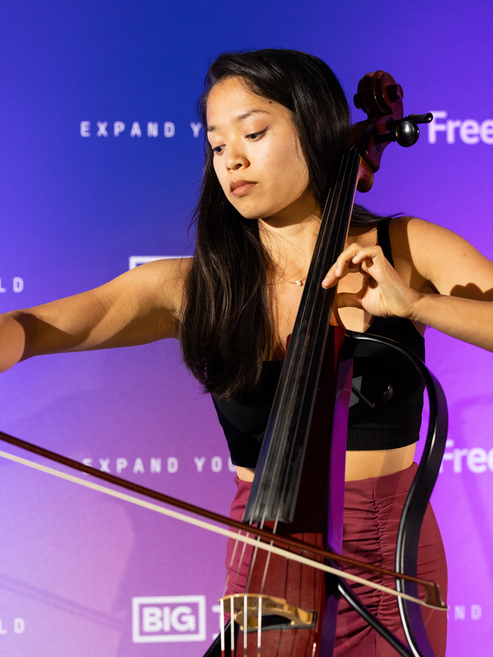 A woman plays an electric cello with a bow on stage in front of a purple backdrop with white text.