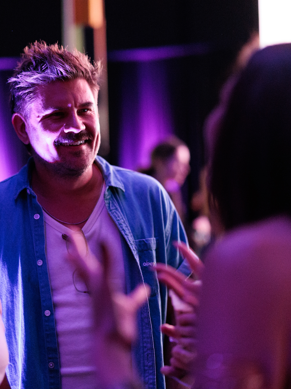 A man in a blue shirt and white t-shirt smiles while talking to someone at an indoor event with purple lighting.