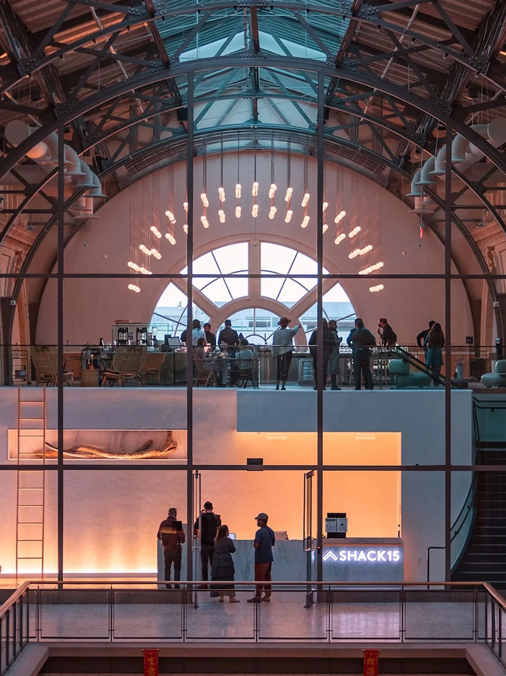 People are gathered on two levels inside a modern building with large arched windows, glass walls, and a bright chandelier; the lower level has a reception desk labeled &ldquo;SHACK15.&rdquo;.