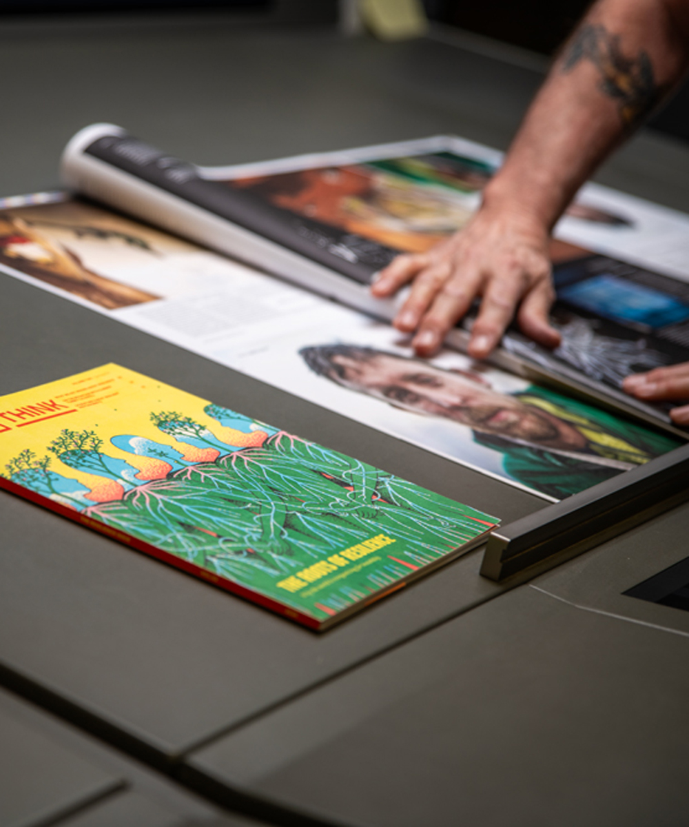 A person’s hands spread open a magazine on a table next to a colorful book featuring illustrated birds and grass on its cover.