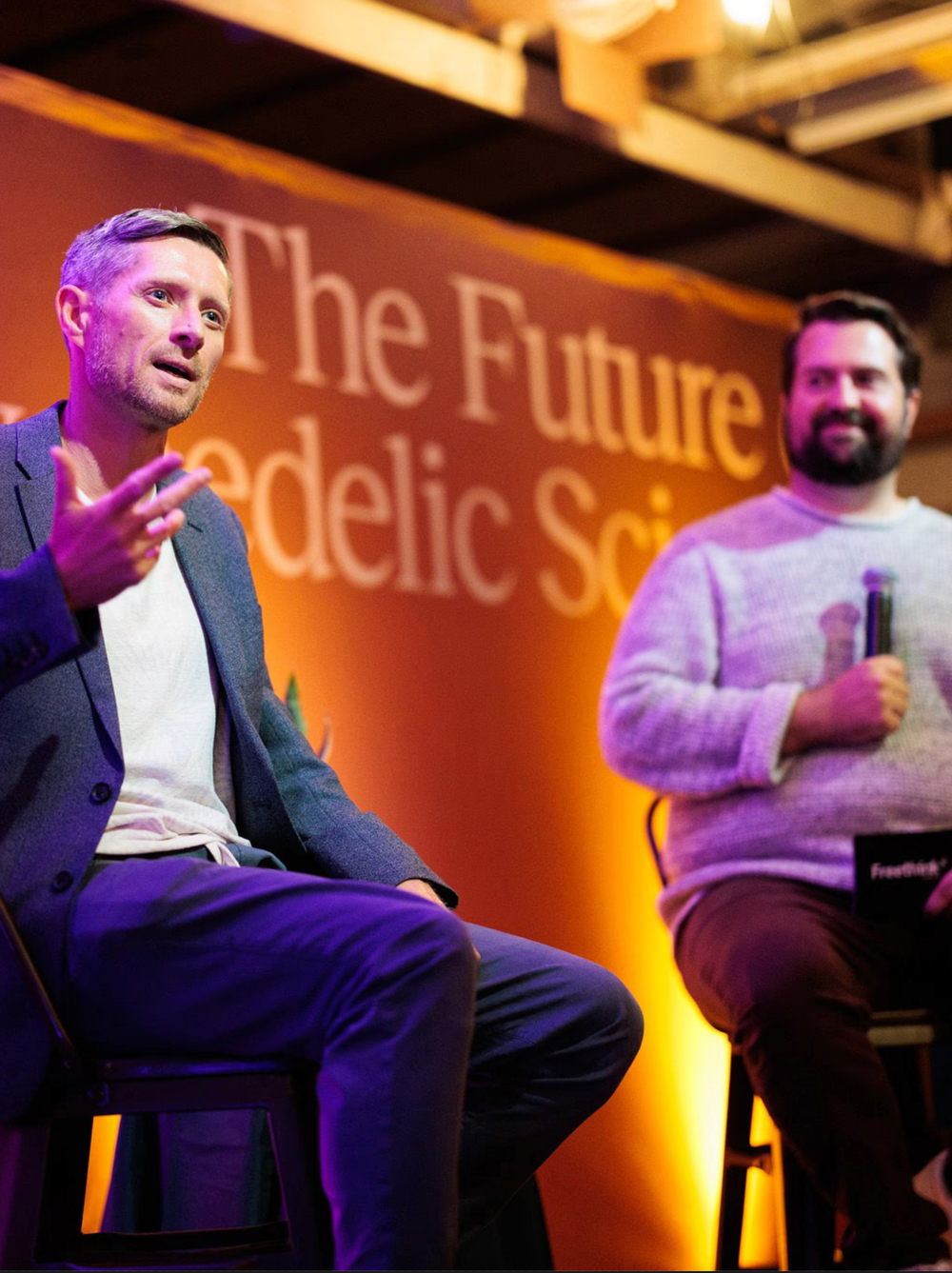 Two men sit on stools speaking at an event with an orange backdrop that reads "The Future of Psychedelic Science." One gestures while talking, the other holds a microphone.