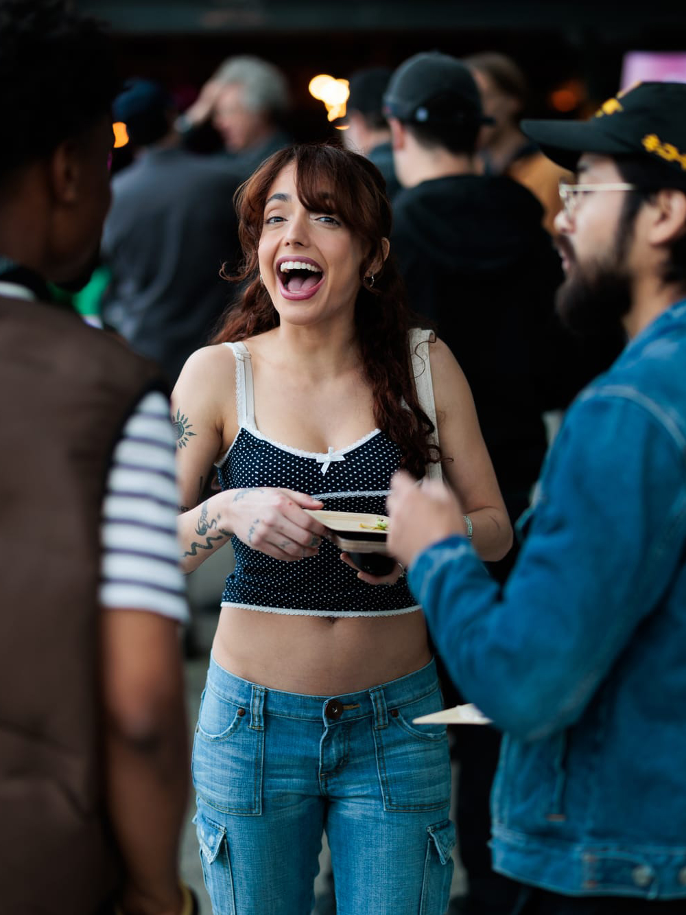 A woman in a crop top and jeans is smiling and talking with two people at an outdoor event; others are blurred in the background.